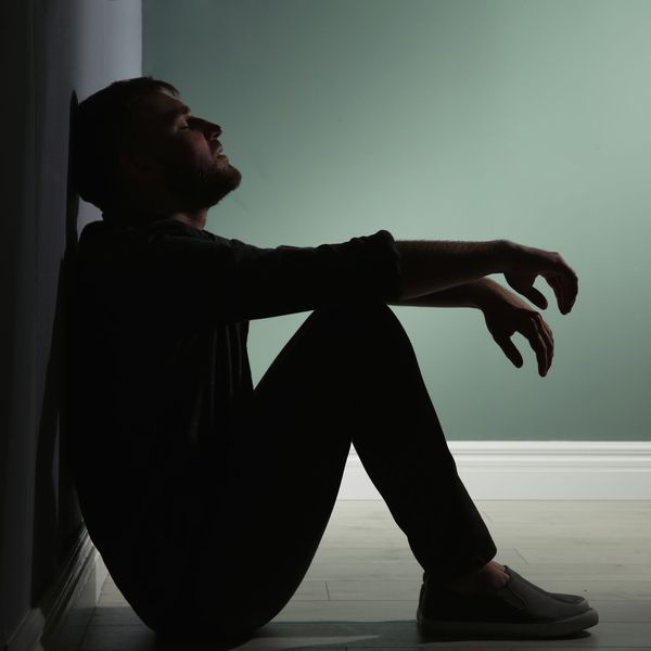 Person sitzt mit angewinkelten Beinen und ausgestreckten Armen an einer Wand in einem dunklen Raum