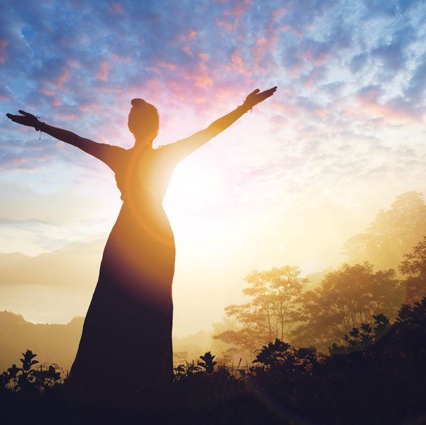 Silhouette einer Person mit ausgebreiteten Armen vor einem Sonnenaufgang über bewaldeten Hügeln